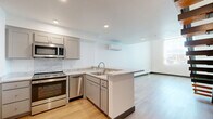This two-story residence offers a thoughtfully designed layout spread across two floors, combining modern finishes with practical functionality. The main level hosts the open-concept living and kitchen area, where light hardwood flooring flows seamlessly throughout. The kitchen is well-appointed with gray shaker-style cabinetry, marble-look countertops, a stainless steel appliance suite including a range, microwave, dishwasher, and refrigerator, and an L-shaped peninsula that doubles as additional prep and serving space. A wall-mounted mini-split unit provides climate control for the open living area, which benefits from recessed lighting and a bright, clean aesthetic. A hallway connects to multiple bathrooms on this level, one of which conveniently houses a stacked washer and dryer unit. The upper floor features the primary bedroom, a generously sized retreat with tall ceilings, a large window that fills the room with natural light, and sliding glass doors opening to an outdoor area. A dedicated closet and additional bathrooms complete the upper level. A staircase with wood treads and metal railings connects both floors. Throughout the residence, crisp white walls, light hardwood flooring, and dark baseboards create a clean, contemporary ambiance.