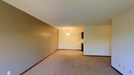 This single-floor residence offers a straightforward, functional layout designed for comfortable everyday living. The main living area is finished with wall-to-wall neutral beige carpeting that extends throughout, providing a warm and cohesive feel underfoot. The walls are painted in a combination of warm taupe and soft off-white tones, creating a calm and inviting atmosphere. A textured popcorn ceiling runs across the space, adding a classic residential character overhead. The living area flows seamlessly into the dining space, which is defined by a pendant light fixture that hangs centrally, casting a warm glow and serving as a visual anchor for the area. Just beyond the dining space, the kitchen comes into view, featuring dark wood cabinetry and what appears to be standard appliances, offering a practical cooking environment. The open-concept connection between the living, dining, and kitchen areas makes the overall floor plan feel spacious and well-suited for both daily routines and casual entertaining. Baseboard trim lines the base of the walls throughout, adding a clean finishing detail. The layout is practical and efficient, making good use of the available space across the single level.