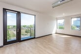 This single-floor loft-style unit showcases a contemporary urban design with an open-concept layout that seamlessly connects the living, dining, and kitchen areas. Light hardwood flooring runs throughout, complementing the crisp white walls and dark baseboard trim. A defining architectural feature is the exposed cylindrical ductwork that traverses the ceiling, lending an industrial character to the space.

The kitchen is thoughtfully equipped with stainless steel appliances, including a full-size refrigerator, electric range, microwave, and dishwasher. Light maple cabinetry with open shelving provides generous storage, while a clean white countertop offers ample prep space. A peninsula counter helps delineate the kitchen from the open living area.

The spacious main living area is bathed in natural light from multiple windows that frame views of the surrounding cityscape and greenery. A large sliding glass door opens to a private balcony with a metal railing, extending the living space outdoors with elevated urban views.

The bathroom features a floating vanity with an undermount sink, a framed mirror, and a walk-in glass shower, all finished with the same hardwood-style flooring found throughout the unit. Recessed lighting and clean fixtures complete the modern, minimalist aesthetic of this well-appointed urban residence.
