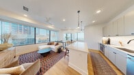 This sophisticated single-floor residence offers a thoughtfully designed open-concept layout with expansive floor-to-ceiling windows that frame striking panoramic city skyline views throughout. Light hardwood flooring flows seamlessly across the entire space, lending warmth and continuity to every room.

The living and dining areas form the heart of the home, with oversized windows wrapping around a dedicated dining nook — an ideal sun-drenched spot to enjoy the urban vista. A ceiling fan and recessed lighting complement the airy atmosphere of the living area.

The kitchen is outfitted with sleek, flat-front cabinetry in a soft neutral tone, a large waterfall-edge quartz island with bar seating, stainless steel appliances including a French door refrigerator, and pendant lighting that adds a refined finishing touch.

The bedroom is a serene retreat, featuring large windows overlooking the cityscape, recessed lighting, and ample space. Double doors lead to a generous walk-in closet with multi-level shelving and hanging rods.

The primary bathroom is elegantly appointed with a long dual-vanity featuring natural wood cabinetry, a white quartz countertop, illuminated mirrors, a glass-enclosed shower with tile surround, and a separate walk-in closet accessible from within.