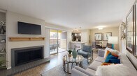 This single-floor residence offers a thoughtfully designed open-concept layout that seamlessly connects the living, dining, and kitchen areas. The living room is a standout space, anchored by a wood-manteled fireplace with built-in shelving on one side and sliding glass doors that open to a private balcony, allowing natural light to fill the room. Light-toned wood-style flooring runs throughout the main living areas, contributing to the space's bright and airy feel.

The kitchen features dark gray cabinetry paired with white quartz-style countertops, stainless steel appliances, and pendant lighting overhead. A breakfast bar connects it to the main living area, making it ideal for entertaining. Adjacent to the kitchen, a dedicated dining nook provides an intimate setting for meals, accented by framed artwork on neutral walls.

The bedroom is a comfortable retreat with plush carpet flooring, a ceiling fan, and direct access to a well-appointed en-suite bathroom. The bathroom includes a full tub and shower combination with classic subway tile surround and wood-look flooring. Throughout the space, a cohesive neutral color palette with teal and warm accent tones creates a modern yet welcoming ambiance.