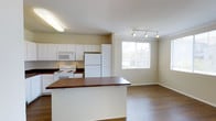 This single-floor space showcases an open-concept layout that seamlessly connects the kitchen and living areas, creating a cohesive and functional environment. The kitchen is outfitted with white cabinetry that extends to the ceiling, providing ample storage, and is complemented by dark countertops that offer a pleasant contrast. A central kitchen island with a rich wood-toned surface serves as a natural dividing point between the kitchen and living areas, offering additional prep space or casual seating. White appliances, including a refrigerator, electric range, and built-in microwave, are neatly integrated into the cabinetry. The flooring throughout is a warm hardwood-style material that adds continuity across both spaces. The living area benefits from a large window that draws in natural light, brightening the neutral-toned walls. Track lighting in the living area provides adjustable illumination, while a fluorescent fixture in the kitchen ensures consistent task lighting. The overall ambiance is clean, bright, and practical, with a straightforward design that prioritizes functionality and open flow.