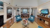 This single-floor community clubhouse and leasing office makes a strong first impression with its dramatic, soaring ceiling heights and classic architectural details. Tall white columns frame the open main lounge area, which is anchored by rich hardwood flooring and a large area rug. The space is furnished with bold teal accent chairs and a sleek glass coffee table, creating a vibrant yet polished atmosphere. Natural light pours in through multiple oversized windows dressed with patterned teal drapery, as well as a set of glass-panel double entry doors that offer views of the surrounding landscape. A secondary seating area features a plush neutral sofa and additional accent chairs, lending a comfortable, residential feel to the space. A rustic wooden console with a mounted flat-screen display serves as an informational station, while a chalkboard welcome sign adds a personal touch. Adjacent to the main lounge, a dedicated private office provides a quiet workspace equipped with a desk and carpeted flooring. A service counter area near the entrance suggests an attendant or concierge function. Throughout, the clean white walls, crown molding, and carefully chosen decor combine to create a professional yet inviting environment.