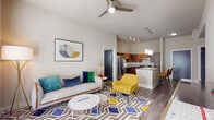 This single-floor residence showcases a thoughtfully designed open-concept layout that seamlessly connects the living, dining, and kitchen areas. The living room features wood-look plank flooring, a ceiling fan, and neutral walls accented by colorful furnishings, including a bold geometric-patterned area rug and vibrant accent chairs. A framed abstract artwork anchors the space visually.

The kitchen is well-appointed with rich walnut-toned cabinetry, granite countertops, a full suite of stainless steel appliances including a gas range, dishwasher, and refrigerator, plus a peninsula island that provides additional prep and seating space. Track lighting and pendant fixtures illuminate the area effectively. The tall ceilings in the kitchen add a sense of openness.

An entry hallway features wood-look flooring and a decorative console, leading to the main living area. French doors allow natural light to filter in from outside.

Both bedrooms are carpeted and comfortably sized, each offering ceiling fans or overhead lighting. One bedroom features a large window for natural light. The bathrooms include granite-topped walnut vanities, tile flooring, and a tiled tub/shower combination with accessibility grab bars, providing both functionality and a clean, modern aesthetic throughout.