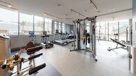 This fitness center is thoughtfully designed across a single level, offering distinct workout zones that cater to a wide range of exercise preferences. The space is flooded with natural light through floor-to-ceiling windows along the perimeter, providing views of the surrounding neighborhood and creating an open, energizing atmosphere throughout.

The cardio zone houses a row of True-brand treadmills and elliptical machines, all equipped with individual digital screens. Adjacent to this area, a dedicated strength training section features a full cable machine system, multiple power racks, weight benches, and a comprehensive dumbbell rack, all set on durable rubberized flooring. Large wall-to-wall mirrors amplify the sense of space and support proper form during workouts.

A separate cycling studio, distinguished by warm wood-look flooring and teal accent walls, contains stationary bikes each paired with personal display screens and is anchored by a large wall-mounted television and built-in wood cabinetry for storage. A functional training area nearby includes stability balls, medicine balls, and resistance accessories organized on a dedicated rack against a geometric-patterned feature wall.

The entry corridor features polished concrete flooring, glass partition walls, and a mounted display screen, creating a clean and modern transition into the facility.