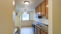 This single-floor space centers around a practical and well-organized kitchen that connects seamlessly to an adjoining living or dining area. The kitchen is lined with warm-toned oak cabinetry, offering generous upper and lower storage. Dark laminate countertops run along the perimeter, complemented by a stainless steel double sink with a curved faucet. A dishwasher is integrated beneath the counter, and a white refrigerator anchors one end of the kitchen. Light tile flooring extends throughout both the kitchen and the adjacent space, creating a cohesive flow between the two areas. A textured ceiling adds a classic residential character overhead, with a flush-mount light fixture illuminating the kitchen. The adjoining room benefits from a large window that brings in natural light, and a ceiling fan with light kit provides both air circulation and additional illumination. White baseboard trim accents the walls, and a window air conditioning unit is visible, suggesting a practical approach to climate control. Overall, the space is functional, well-lit, and comfortably laid out for everyday living.