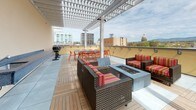 The fifth-floor patio is a well-appointed outdoor terrace designed for both relaxation and social gatherings. A prominent white pergola spans a portion of the space, providing partial shade while maintaining an open-air ambiance and framing sweeping views of the surrounding cityscape, including a notable domed capitol building, as well as distant mountain ridgelines.

The patio features a combination of large-format tile and warm wood decking underfoot, creating a thoughtful contrast of materials throughout. A central fire pit table anchors a comfortable lounge area furnished with wicker seating accented by bold striped cushions, ideal for casual conversation or evening gatherings. Nearby, a dining table with chairs accommodates group meals, and a grill station supports outdoor cooking.

Along one wall, a granite countertop with an integrated sink serves as a functional outdoor utility station, complemented by a stainless steel waste bin. Large planters with greenery line the perimeter, adding natural texture to the environment. The overall aesthetic is clean and modern, with the expansive open sky and panoramic views serving as the patio's defining feature.
