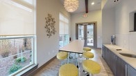 The Cyber Cafe is a thoughtfully designed, single-floor communal workspace that blends functional amenities with contemporary style. The space is anchored by a long, sleek white communal table surrounded by adjustable yellow upholstered stools, providing a collaborative setting for work or socializing. A striking sculptural pendant light fixture serves as a focal point overhead, complementing the exposed dark wood ceiling beams that add architectural character.

Along one wall, a continuous run of dark wood cabinetry with light countertops houses a built-in sink and is topped by a wall-mounted flat-screen television, making the space versatile for both productivity and entertainment. A textured tile backsplash adds visual interest behind the cabinetry.

Natural light flows generously through multiple large windows fitted with roller shades, offering views of the surrounding outdoor areas. A set of double glass doors at one end provides direct access to the exterior courtyard. Light wood-toned flooring runs throughout, enhancing the bright, airy ambiance. Decorative wall art, including a sculptural metal piece and a graphic canvas, adds personality to the neutral-toned walls, creating an inviting and modern atmosphere throughout the space.