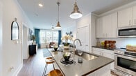 This thoughtfully designed single-story residence offers a cohesive open-concept layout that efficiently connects the kitchen, dining, and living areas. The kitchen serves as a central hub, featuring white shaker-style cabinetry, dark countertops, a stainless steel appliance suite including a range, microwave, and refrigerator, a white subway tile backsplash, and a large island with seating for two—illuminated by brushed brass pendant lights. Hardwood-style flooring runs throughout the main living areas, adding warmth and continuity.

The living room is bright and generously proportioned, benefiting from multiple large windows dressed with teal drapes that invite natural light while offering views to the exterior. Crown molding and a ceiling fan add architectural detail to the space.

The primary bedroom accommodates a full bedroom suite and features a prominent window with blinds and floor-length drapes, providing natural light and outdoor views. The full bathroom continues the unit's clean aesthetic with white cabinetry, a dark countertop vanity, a large mirror, wood-look flooring, and crisp white walls. Two hallways connect the spaces, contributing to an efficient and functional overall floor plan.