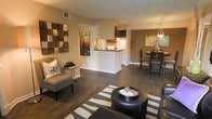 This single-floor residence offers a thoughtfully designed open-concept layout that seamlessly connects the living, dining, and kitchen areas into one cohesive and functional space. Rich, dark hardwood flooring flows throughout, grounding the warm, neutral color palette that defines the interior's inviting ambiance.

The living area is generously proportioned, providing ample room for comfortable seating arrangements. A decorative geometric wall panel serves as a bold focal point, complemented by vibrant abstract artwork that adds color and personality to the space.

The kitchen features a raised breakfast bar that acts as a natural divider between the living and cooking areas while maintaining the open feel. Dark cabinetry contrasts with light countertops and white appliances, creating a classic and practical culinary space.

Adjacent to the kitchen, the dining area is anchored by a stylish pendant light that casts warm illumination over the dining table and chairs. A large piece of colorful artwork adorns the accent wall, further enhancing the room's character.

Together, these spaces create a harmonious and well-lit interior that balances everyday functionality with aesthetic appeal, making it an ideal setting for both relaxed living and entertaining.