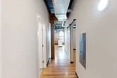 Located at 1 Websters Landing in downtown Syracuse, NY, this industrial-style loft showcases a striking blend of historic character and contemporary design. The space occupies a single floor and includes a living room, hallway, bathroom, and closet.

The hallway sets the tone immediately, featuring warm hardwood flooring, exposed ductwork overhead, a painted steel ceiling structure, and a sliding barn door — all hallmarks of a thoughtful industrial conversion. Reclaimed wood posts and brick accent walls add warmth and authenticity throughout.

The living room is an open, light-filled space anchored by a rustic timber column and complemented by soaring exposed ceilings. Two dramatic arched windows frame sweeping views of the Syracuse cityscape and waterway, allowing generous natural light to pour in across the hardwood floors.

The kitchen area features white cabinetry, a granite-topped island, and stainless steel appliances, seamlessly integrated into the open layout while maintaining the industrial aesthetic of the space.

The bathroom is clean and modern, finished with stone-look tile flooring, tile wainscoting, a white vanity with granite countertop, and a glass-enclosed shower area with accessibility features.

Overall, this space delivers a refined urban loft experience, balancing raw architectural character with polished, move-in-ready modern amenities.