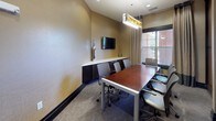 This single-floor conference room offers a polished, professional environment designed for productive meetings. At its center sits a rich cherry-toned rectangular conference table surrounded by ergonomic, adjustable office chairs, comfortably accommodating a small group. A modern pendant light fixture with warm amber glass detailing hangs overhead, providing focused illumination and serving as a distinctive design element.

One wall features a sleek, built-in credenza with dark cabinetry and a white countertop, above which a wall-mounted flat-screen television is positioned for presentations and video conferencing. Natural light filters into the room through a window fitted with horizontal blinds and floor-length drapes in neutral tones, offering views to the exterior.

On the opposite side, a striking frosted glass partition with dark framing adds a layer of sophistication, while a pair of framed abstract artworks in earth tones — browns, creams, and muted reds — contribute a curated, contemporary aesthetic. Neutral-toned textured walls and carpeted flooring complete the refined interior. A doorway at the rear connects the space to an adjoining corridor, ensuring convenient access.