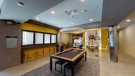 Located at 1001 W Rosedale St in Fort Worth, Texas, this single-floor amenity space blends contemporary design with locally inspired character. The layout is divided into distinct functional zones, each thoughtfully designed to encourage both socializing and relaxation.

The game room serves as a central focal point, anchored by a sleek billiards table beneath a striking circular pendant light. Bold yellow accent walls energize the space, while a typography-style mural celebrating Fort Worth culture adds a strong sense of local identity. Bar-height stools line one wall, and an adjacent dining area with high-top tables and pendant lighting provides additional gathering space.

Nearby, a compact kitchenette features warm gold cabinetry, a dark stone countertop, a built-in microwave, and a pod-style coffee station set against a textured three-dimensional hexagonal tile wall — a refined architectural detail that elevates the utilitarian space.

A separate lounge area offers a more relaxed atmosphere, furnished with plush leather seating arranged around a glass coffee table atop a richly patterned area rug. A wall-mounted flat-screen television is framed by subtly lit tile paneling. Floor-to-ceiling glass doors open to an outdoor area, allowing natural light to filter in. Curated abstract artwork displayed in the hallways completes the polished, gallery-like ambiance throughout.