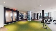 Located at 100 Hale Ave in White Plains, NY, this well-appointed fitness center offers a thoughtfully designed workout environment across a single, open floor. The space is organized into distinct functional zones that cater to a wide range of training styles and fitness goals.

A standout design element is the striking floor-to-ceiling nature mural depicting a misty forest, which adds an atmospheric and visually dynamic focal point throughout the gym. Bright white ceilings with recessed lighting keep the space feeling open and well-illuminated, while large windows along one wall invite natural light and provide views of the surrounding neighborhood.

The central training area features vibrant artificial turf flooring, ideal for functional movement, stretching, and agility drills. Adjacent to this zone is a dedicated strength and conditioning section equipped with a multi-functional cable machine, a full dumbbell rack with an adjustable bench, and a rowing machine. A separate cardio zone houses multiple treadmills and an elliptical trainer, all positioned to face the natural light from the windows.

A distinct boxing and high-intensity training alcove includes a punching bag, resistance bands, and interactive display screens for guided workouts. Dark accent columns and panels provide modern contrast against the clean white walls, contributing to a sleek, contemporary aesthetic that makes this fitness facility both functional and visually engaging.