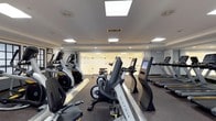 This fitness center is a thoughtfully designed, single-floor facility offering a complete workout environment for cardio enthusiasts and strength trainers alike. The space is divided into two well-defined zones, each catering to different aspects of physical fitness.

The cardio area is outfitted with a lineup of modern Matrix-brand machines, including treadmills, upright and recumbent bikes, and elliptical cross-trainers. Each machine features an integrated digital display, providing users with real-time performance data. Skylights and recessed panel lighting illuminate the area brightly, creating an energizing atmosphere. Large glass panels along one wall add openness to the space and allow visibility between areas.

The strength training zone is equally impressive, anchored by a central multi-functional cable machine that enables a wide variety of resistance exercises. Surrounding it are several weight benches and dedicated resistance machines with padded orange seating, catering to targeted muscle training. A well-stocked dumbbell rack lines one wall, offering a full range of free weights for versatile training options.

The facility features rubber flooring throughout for safety and durability, smooth neutral-toned walls, and a clean, modern aesthetic. Recessed and flush-mounted ceiling lights ensure consistent illumination across the entire space, making it a practical and inviting environment for daily fitness routines.