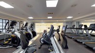 This fitness center is a thoughtfully designed, single-floor facility offering a complete workout environment for cardio enthusiasts and strength trainers alike. The space is divided into two well-defined zones, each catering to different aspects of physical fitness.

The cardio area is outfitted with a lineup of modern Matrix-brand machines, including treadmills, upright and recumbent bikes, and elliptical cross-trainers. Each machine features an integrated digital display, providing users with real-time performance data. Skylights and recessed panel lighting illuminate the area brightly, creating an energizing atmosphere. Large glass panels along one wall add openness to the space and allow visibility between areas.

The strength training zone is equally impressive, anchored by a central multi-functional cable machine that enables a wide variety of resistance exercises. Surrounding it are several weight benches and dedicated resistance machines with padded orange seating, catering to targeted muscle training. A well-stocked dumbbell rack lines one wall, offering a full range of free weights for versatile training options.

The facility features rubber flooring throughout for safety and durability, smooth neutral-toned walls, and a clean, modern aesthetic. Recessed and flush-mounted ceiling lights ensure consistent illumination across the entire space, making it a practical and inviting environment for daily fitness routines.