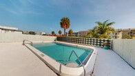 This single-floor space centers around a well-maintained outdoor swimming pool that serves as its defining feature. The rectangular pool is finished in a clean, light-colored surround that reflects the abundant natural sunlight, with stainless steel entry rails providing easy access into the clear turquoise water. A striking decorative accent wall runs along one side of the pool area, adorned with a classic diamond-pattern checkered tile design in green and white tones, lending a retro, resort-style character to the space. The pool deck is generously sized, offering ample room around the water's edge. Tall, mature palm trees rise above the perimeter walls, creating a tropical ambiance and providing a sense of seclusion while framing views of the surrounding neighborhood and open blue sky above. Potted plants accent the corners, adding greenery and a welcoming touch to the area. The overall atmosphere is bright, open, and relaxed, evoking a classic Florida leisure setting suited for outdoor recreation and enjoyment.