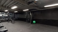 Located at 253 Academy St in Jersey City, NJ, this single-floor fitness facility presents a purposefully designed workout environment defined by a consistent industrial aesthetic. The space is finished in an all-black color scheme throughout, with dark-painted walls, exposed metal ceilings, and visible ductwork and piping overhead, lending the facility a raw, utilitarian character. Flush-mounted fluorescent panel lighting provides even illumination across the open floor plan.

The flooring throughout is covered in interlocking rubber mats, offering both durability and traction suitable for high-intensity training. The space is divided into two functional zones. One area is dedicated to strength training and includes a cable machine, a Rogue power rack, a flat bench, and a barbell setup, with floor-to-ceiling mirrors lining one wall to maximize visual depth and allow for form monitoring. A stability ball and additional free weights complement the setup.

The second zone is oriented toward cardio and open-floor training, housing Spirit treadmills and additional cardio equipment. A digital fitness station is mounted near the exit door in this area. The space also includes a ceiling-mounted HVAC unit and a clearly marked emergency exit, ensuring comfort and safety. Overall, the facility delivers a focused, no-frills training environment.