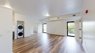Located at 1116 S Walton Blvd in Bentonville, Arkansas, this single-floor unit offers a clean, modern design with a practical and open layout. The main living area is expansive and filled with natural light, courtesy of two black-framed glass doors that open onto a shared balcony, offering outdoor views of the surrounding area. Durable wood-look flooring runs throughout the main living space, complementing the bright white walls and creating a warm, contemporary atmosphere.

The kitchen is well-appointed with two-tone cabinetry — white upper cabinets paired with teal lower cabinets — white countertops, and a full suite of stainless steel appliances, including a refrigerator, dishwasher, range, and microwave. A stacked washer and dryer unit is conveniently positioned adjacent to the kitchen.

The bedrooms feature neutral carpet flooring, natural light from windows with blinds, and closet storage. The bathroom is notably spacious and accessibility-conscious, featuring a floating teal vanity, large mirror, tile flooring, and a generously sized roll-in shower with a handheld showerhead and grab bar, finished in white subway tile.