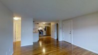 This single-floor residence offers a practical and well-maintained open-concept layout that seamlessly connects the living area and kitchen. The warm-toned hardwood floors extend throughout the main living space, lending a sense of continuity and warmth to the interior.

The kitchen is efficiently designed with white cabinetry, light countertops, and a full suite of stainless steel appliances, including a gas range and a refrigerator. The cabinetry provides ample storage, while the layout allows for easy movement and functionality during meal preparation. A sink is positioned along the counter, completing the workspace.

The open living area is generous in scale, offering flexible space for a variety of furniture arrangements. White walls and a textured ceiling keep the atmosphere clean and neutral, allowing the natural tones of the hardwood floors to stand out. A solid white entry door is visible from the main living area, providing a clear sense of the unit's entry point.

A hallway leads from the main living space toward additional areas of the unit, suggesting further rooms beyond the open plan. Overall, the space presents a clean, functional, and inviting environment with classic finishes that offer a timeless and adaptable aesthetic.