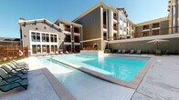 This outdoor pool area is a standout amenity within a well-appointed multi-story residential community. The centerpiece is a generously sized swimming pool with crystal-clear blue water, finished with clean white tile surrounds and elegant edge detailing that gives it a modern, resort-like aesthetic. The pool deck is spacious and open, offering ample room for lounging, with multiple chaise lounge chairs arranged along the perimeter to take full advantage of the sunny, open-sky setting.

The surrounding building architecture features a combination of horizontal lap siding in muted earth tones — sage green and warm gray — accented by deep red-brown trim and private balconies that overlook the pool area. Lush tropical plantings, including palm trees and colorful flowering plants, add a vibrant touch to the landscaping throughout.

A covered outdoor corridor runs along the building's ground level, providing shaded access between building entrances. This covered walkway features a ceiling fan, recessed lighting, and speckled concrete flooring, creating a comfortable transitional space even in warm weather. Entryways are marked by solid double doors with warm wood-toned finishes set against the sage lap-siding exterior. The overall environment is bright, clean, and thoughtfully designed to provide a relaxing outdoor retreat.