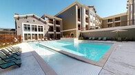 This outdoor pool area is a standout amenity within a well-appointed multi-story residential community. The centerpiece is a generously sized swimming pool with crystal-clear blue water, finished with clean white tile surrounds and elegant edge detailing that gives it a modern, resort-like aesthetic. The pool deck is spacious and open, offering ample room for lounging, with multiple chaise lounge chairs arranged along the perimeter to take full advantage of the sunny, open-sky setting.

The surrounding building architecture features a combination of horizontal lap siding in muted earth tones — sage green and warm gray — accented by deep red-brown trim and private balconies that overlook the pool area. Lush tropical plantings, including palm trees and colorful flowering plants, add a vibrant touch to the landscaping throughout.

A covered outdoor corridor runs along the building's ground level, providing shaded access between building entrances. This covered walkway features a ceiling fan, recessed lighting, and speckled concrete flooring, creating a comfortable transitional space even in warm weather. Entryways are marked by solid double doors with warm wood-toned finishes set against the sage lap-siding exterior. The overall environment is bright, clean, and thoughtfully designed to provide a relaxing outdoor retreat.