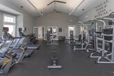 Located at 980 Walther Blvd in Lawrenceville, GA, this single-floor fitness center is a comprehensively outfitted facility designed to support a full range of workout routines. The space is defined by its striking vaulted ceilings, which lend an open and airy atmosphere throughout. Large, multi-pane windows line the walls, allowing abundant natural light to flood the interior and offering views of the outdoor surroundings, including what appears to be a nearby pool area.

The facility is divided into distinct workout zones. The cardio section features multiple treadmills, elliptical trainers, stationary bikes, and a rowing machine, all arranged along the windowed walls to maximize the sense of space and natural light. Ceiling fans overhead contribute to a comfortable exercise environment. The strength training area is equipped with a robust selection of cable machines, weight benches, multi-station strength machines, and a well-stocked dumbbell rack. Full-length mirrors are mounted along the walls, enhancing the open feel of the space and providing functional visibility during workouts.

The interior is finished with dark rubber flooring throughout, offering durability and safety underfoot. Neutral gray walls provide a clean, modern backdrop, complemented by a motivational "Sweat, Smile and Repeat" wall decal. The overall ambiance is bright, professional, and welcoming for fitness enthusiasts of all levels.