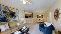 This single-floor residence offers an open-concept layout that seamlessly connects the living, dining, and kitchen areas into one cohesive and functional space. The living area is anchored by plush carpeting, crown molding, and a ceiling fan, creating a comfortable and inviting atmosphere. A large abstract wall painting adds a bold artistic touch, while ample natural light enhances the warm, neutral color palette throughout.

The kitchen features warm wood cabinetry, black appliances, and a raised breakfast bar that serves as a natural divider between the kitchen and the living space. This semi-open design allows for easy interaction between rooms while maintaining a defined cooking area.

Adjacent to the living room, the dining area is highlighted by an elegant chandelier that adds a refined touch to the space. The dining setup comfortably accommodates a full table and chairs, making it well-suited for both everyday meals and entertaining guests.

Throughout the space, consistent crown molding, neutral wall tones, and soft carpeting contribute to a cohesive and polished aesthetic. The overall ambiance is warm and welcoming, with thoughtful design details that balance everyday practicality with understated elegance.