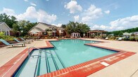 This community space features a elegantly appointed clubhouse lobby and a well-designed outdoor pool area. The lobby makes a strong first impression with its refined architectural details, including tall decorative columns finished in a brushed silver tone, raised panel wainscoting, and a contrasting tile floor of polished black marble and light neutral tile arranged in a geometric pattern. A striking contemporary chandelier serves as the centerpiece overhead, casting warm light across the space. Large windows dressed with textured drape panels allow ample natural light to filter in, enhancing the airy ambiance. A glass-topped round table with ornate silver décor anchors the center of the lobby, while bold abstract artwork adorns the walls. French doors with glass pane inserts lead to an adjacent office or leasing area, and a bench provides a convenient waiting spot near the entrance.

The outdoor pool area offers a generously sized swimming pool with a classic brick-tile surround, complemented by a broad concrete deck furnished with lounge chairs and shaded by a market umbrella. A pergola structure at the rear of the pool deck provides additional shade and a defined outdoor gathering space. Mature trees and landscaping frame the area, and the surrounding residential buildings are visible in the background, reinforcing the sense of a cohesive community environment.