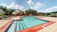 This community space features a elegantly appointed clubhouse lobby and a well-designed outdoor pool area. The lobby makes a strong first impression with its refined architectural details, including tall decorative columns finished in a brushed silver tone, raised panel wainscoting, and a contrasting tile floor of polished black marble and light neutral tile arranged in a geometric pattern. A striking contemporary chandelier serves as the centerpiece overhead, casting warm light across the space. Large windows dressed with textured drape panels allow ample natural light to filter in, enhancing the airy ambiance. A glass-topped round table with ornate silver décor anchors the center of the lobby, while bold abstract artwork adorns the walls. French doors with glass pane inserts lead to an adjacent office or leasing area, and a bench provides a convenient waiting spot near the entrance.

The outdoor pool area offers a generously sized swimming pool with a classic brick-tile surround, complemented by a broad concrete deck furnished with lounge chairs and shaded by a market umbrella. A pergola structure at the rear of the pool deck provides additional shade and a defined outdoor gathering space. Mature trees and landscaping frame the area, and the surrounding residential buildings are visible in the background, reinforcing the sense of a cohesive community environment.