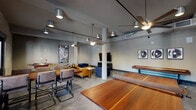 This single-story club room in New Orleans, LA, masterfully combines industrial character with contemporary comfort. A hallway entrance lined with exposed brick walls and polished concrete floors sets the tone, leading into a spacious, open main area defined by structural columns, ceiling fans, and track lighting overhead.

The lounge zone anchors one side of the room, furnished with a rich amber leather sofa, a navy blue armchair, and a large flat-screen television mounted above a media console—ideal for relaxed gatherings. Original artwork adorns the walls throughout, adding a curated, creative atmosphere. A foosball table and a regulation ping-pong table provide entertainment options, while a shuffleboard table extends the game room experience further.

A dedicated wet bar area features dark wood cabinetry, a sink, and under-counter refrigeration, making the space well-equipped for hosting. A large communal dining table surrounded by upholstered cantilever chairs sits adjacent to French doors and single-panel doors that open directly to an outdoor patio, flooding the area with natural light and offering views of lush exterior greenery. The exposed brick continues in this dining nook, reinforcing the building's historic New Orleans character throughout.
