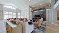 This expansive single-floor clubhouse-style space is designed to impress, combining architectural grandeur with thoughtfully curated interiors. The main lounge area commands attention with its dramatic double-height ceilings, supported by structural columns and overlooked by an upper mezzanine level with wrought-iron railings. A floor-to-ceiling tiled feature wall anchors the room, housing a large flat-screen television flanked by open shelving. Seating arrangements feature tan leather wingback chairs, bold cobalt blue ottomans, and plush sofas positioned atop a richly patterned area rug, all set on geometric-patterned tile flooring.

Adjacent to the lounge, a bright dining room benefits from towering windows dressed in sheer linen curtains, offering views of an outdoor pool and surrounding grounds. Dark wood tables paired with green-cushioned chairs fill this sunlit room, which also features a wall-mounted television and decorative mirror gallery wall.

The fully equipped kitchen stands out with its striking two-tone cabinetry—vibrant cobalt blue lower cabinets contrasted with crisp white uppers—complemented by quartz countertops, stainless steel appliances, a wine cooler, a central island with bar seating, and pendant lighting overhead. Throughout the space, geometric tile flooring and recessed lighting create a cohesive, polished aesthetic.