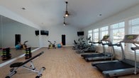 Located at 3151 Boulevard De Sevilla in Foley, Alabama, this fitness center is a thoughtfully designed, single-floor facility built to support a well-rounded workout routine. The space features vaulted ceilings with ceiling fans that promote airflow and contribute to an open, airy atmosphere. Light wood-style flooring runs throughout, lending a clean and modern aesthetic.

The cardio zone is generously equipped with multiple treadmills, elliptical cross-trainers, and a stationary cycling bike, all positioned along a bank of large windows fitted with plantation-style blinds. These windows flood the space with natural light and offer views of the surrounding outdoor area, including what appears to be a pool and neighboring structures.

A dedicated strength and flexibility area includes a full rack of color-coded dumbbells, a weight bench, stability balls, and a wall-mounted punching bag — providing ample options for resistance and functional training. Full-length mirrors line one wall, visually expanding the space and aiding in form correction during exercise.

Multiple wall-mounted flat-screen televisions are positioned throughout the room, ensuring entertainment from virtually any workout station. An adjacent room with glass-paned French doors is visible, offering additional community space. The overall design is bright, clean, and purposefully organized to accommodate a variety of fitness needs.
