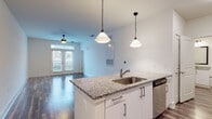 This single-floor unit at The Oliver offers a thoughtfully designed open-concept layout that seamlessly connects the kitchen and living areas. The kitchen serves as a central feature, anchored by a peninsula island topped with granite countertops and an undermount stainless steel sink. Pendant lights hang above the island, providing focused task lighting while contributing to the space's modern aesthetic. White cabinetry with brushed nickel hardware lines the kitchen walls, complemented by a subway tile backsplash and a full suite of stainless steel appliances, including a refrigerator, range, microwave, and dishwasher.

The living area extends from the kitchen, finished with rich, dark hardwood-style flooring throughout and a ceiling fan overhead. French doors at the far end of the living space invite natural light and provide access to an outdoor area. The bedroom features plush carpeting and a ceiling fan, offering a comfortable retreat. The bathroom includes a tub and shower combination with classic white square tile surround, granite-topped vanity, and matching hardwood-style flooring, maintaining a cohesive design throughout the unit.