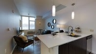This contemporary single-floor residence showcases a thoughtfully designed open-concept layout that integrates the kitchen, dining, and living areas into one cohesive and functional space. The kitchen is a standout feature, equipped with a generous white quartz waterfall island that serves as both a prep surface and a casual dining counter. Dark cabinetry provides a striking contrast to the bright countertops, while sleek pendant lighting adds a modern, refined touch overhead. The kitchen flows effortlessly into the living area, which is bathed in natural light courtesy of expansive floor-to-ceiling windows along the exterior wall. The living space is anchored by hardwood-style flooring and is accented with contemporary furnishings, including a tufted sofa, a leather accent chair, and a layered geometric area rug. A cluster of round decorative mirrors adorns one wall, adding depth and visual interest to the open floor plan. The dining area sits adjacent to the windows, offering a bright spot for meals. Throughout the space, a neutral color palette of whites, grays, and warm wood tones creates a clean, modern ambiance that feels both inviting and sophisticated.