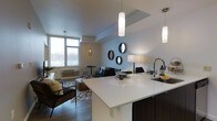 This contemporary single-floor residence showcases a thoughtfully designed open-concept layout that integrates the kitchen, dining, and living areas into one cohesive and functional space. The kitchen is a standout feature, equipped with a generous white quartz waterfall island that serves as both a prep surface and a casual dining counter. Dark cabinetry provides a striking contrast to the bright countertops, while sleek pendant lighting adds a modern, refined touch overhead. The kitchen flows effortlessly into the living area, which is bathed in natural light courtesy of expansive floor-to-ceiling windows along the exterior wall. The living space is anchored by hardwood-style flooring and is accented with contemporary furnishings, including a tufted sofa, a leather accent chair, and a layered geometric area rug. A cluster of round decorative mirrors adorns one wall, adding depth and visual interest to the open floor plan. The dining area sits adjacent to the windows, offering a bright spot for meals. Throughout the space, a neutral color palette of whites, grays, and warm wood tones creates a clean, modern ambiance that feels both inviting and sophisticated.