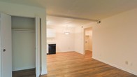 This single-floor space offers a clean, open layout designed for comfortable everyday living. Warm hardwood flooring flows throughout, tying the living, dining, and kitchen areas together in a cohesive and inviting manner. The kitchen is outfitted with white cabinetry, a black dishwasher, and is illuminated by a classic pendant light that hangs above, adding a subtle decorative touch. Crisp white walls and smooth ceilings reflect light generously, creating a bright and airy atmosphere throughout. A bi-fold closet door near the entryway provides convenient storage. Ceiling vents are positioned thoughtfully for efficient climate control. Doorways connect to additional rooms, hinting at further functional spaces beyond the main living area. The overall ambiance is clean, neutral, and move-in ready, offering a versatile canvas well-suited to a variety of interior styles. The open flow between spaces makes the layout feel expansive and practical, ideal for both relaxed living and everyday functionality.