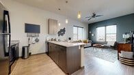 This well-appointed single-floor residence offers a thoughtfully designed open-concept layout that seamlessly connects the kitchen and living areas. The kitchen is a standout feature, showcasing a large peninsula with granite countertops and bar seating, two-tone cabinetry with light upper and dark lower cabinets, black appliances including a full-size refrigerator, range, and microwave, and stylish pendant lighting overhead. Light wood-style flooring runs throughout the main living areas, enhancing the bright and airy feel of the space. The living room is generously sized, featuring large windows on two walls that invite abundant natural light and outdoor views. A teal accent wall adds a bold design touch, while a ceiling fan provides additional comfort. Two bedrooms offer carpeted floors and natural light through large windows, providing a comfortable and restful retreat. The bathroom is finished with large-format gray tile surrounding a full soaking tub and shower, complemented by dark upper cabinetry for added storage. Throughout the residence, the design strikes a balance between modern finishes and warm, inviting tones, creating a cohesive and functional living environment.