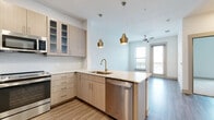 This single-floor unit showcases a contemporary design with a cohesive, modern aesthetic carried throughout. The open-concept kitchen is a central highlight, featuring light wood cabinetry with glass-front upper cabinets, stone-look countertops, and a textured tile backsplash. Stainless steel appliances — including a range, microwave, refrigerator, and dishwasher — complement the space, while gold-tone pendant lights and a matching faucet add warmth and visual interest. A peninsula with extended counter space provides additional prep and seating areas, seamlessly connecting the kitchen to the living area.

The living area is bright and open, with wood-look flooring, recessed lighting, a ceiling fan, and access to an outdoor balcony through glass doors that invite natural light. The bedroom is finished with plush carpet, a ceiling fan, and a window offering outside views, creating a calm and comfortable atmosphere.

The bathroom is well-proportioned, featuring wood-look flooring, a double-sink vanity with stone countertops, light wood cabinetry, a large mirror, open shelving, and a tub-shower combination with tile surround. The overall design is clean, functional, and cohesive.