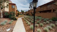 This ground-floor unit, designated as #123, is part of a multi-building residential complex that showcases a warm, Spanish-influenced architectural style. The buildings feature textured stucco exteriors in earth tones, red-tiled rooflines, and wood-accented balconies and fencing that lend the community a cohesive, inviting aesthetic. The unit is accessed via a concrete walkway and is immediately identifiable by its bold red entry door, which stands out against the cream-colored stucco facade. A sliding window adjacent to the entrance allows for natural light and ventilation. Overhead, a black metal staircase leads to upper-level units, adding a structural element to the exterior composition. The surrounding common areas are thoughtfully landscaped with drought-tolerant ornamental grasses, mulched garden beds, mature trees, and classic black lantern-style light posts that line the pedestrian pathways. These walkways wind through the complex, connecting buildings and creating a pleasant, park-like environment. Views from the property extend toward a distant mountain range, offering a scenic natural backdrop. The overall ambiance of the complex is calm and well-kept, reflecting a community designed for comfortable, everyday living with attention to both functionality and curb appeal.