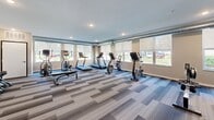 Located at 704 First Dr NW in Austin, Minnesota, this single-floor fitness center offers a well-equipped and thoughtfully designed workout space. The room features a modern aesthetic, anchored by a patterned accent wall with an integrated flat-screen TV, creating a dynamic focal point within the space. The flooring throughout consists of multi-toned modular carpet tiles in varying shades of gray and blue, adding a contemporary, clean look while providing underfoot comfort during workouts.

Natural light is a defining characteristic of this fitness center, with a row of large windows fitted with roller shades lining one wall and offering outdoor views of the surrounding greenery. Exposed ductwork runs along the upper perimeter, contributing an industrial design element that complements the overall modern aesthetic.

The cardio zone is well-appointed, featuring multiple treadmills, upright stationary bikes, and an elliptical trainer positioned to take advantage of the natural light and outdoor views. On the opposite end of the room, a dedicated strength training area includes a multi-station cable machine, a full rack of dumbbells ranging across various weights, additional adjustable dumbbells, and a workout bench. Stability balls and supplementary equipment are neatly organized near the accent wall. Recessed ceiling lighting ensures the space remains bright and functional throughout all hours.