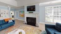 Located at 1600 Jasmine Cove Circle in Simpsonville, South Carolina, this single-floor community clubhouse offers a welcoming and well-appointed gathering space designed for comfort and socializing. The main lounge area is anchored by a classic white-mantled fireplace with a mounted flat-screen television above, creating a natural focal point for the room. Light blue walls, crisp white crown molding, and light wood-look flooring run throughout, lending the space a clean, airy aesthetic. A vaulted ceiling with a ceiling fan adds a sense of openness and enhances airflow across the generously sized lounge. Built-in white cabinetry and shelving along one wall provide both display and storage functionality, complementing the room's polished, cohesive design. Large windows along the perimeter flood the interior with abundant natural light, while a set of glass-paned French doors opens directly to an outdoor pool area, seamlessly connecting the interior to the exterior. Colorful area rugs and coordinated blue upholstered seating arrangements create inviting conversation areas throughout the space. A secondary seating nook near the pool-facing doors offers a bright, relaxed setting ideal for casual gatherings. The overall ambiance is cheerful, bright, and thoughtfully designed to serve as a versatile community hub.