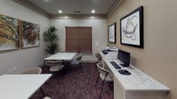 This business center is a well-appointed, single-floor facility designed to support productivity and collaborative work. The space is thoughtfully organized into two functional zones: a dedicated workstation area and an open meeting area.

The workstation area features a granite-topped counter desk equipped with a desktop computer, keyboard, mouse, and a multifunction printer/scanner, providing all the essentials for document management and digital tasks. A tall potted plant beside a window with brown horizontal blinds adds a refreshing, natural touch to this side of the room.

The meeting area accommodates multiple white square tables paired with comfortable cushioned chairs, offering ample space for group discussions or individual work. The patterned deep-burgundy carpet anchors the room with a warm, professional aesthetic.

Décor throughout the space includes framed abstract and nature-inspired artwork, enhancing the visual appeal of the neutral-toned walls. Recessed lighting provides consistent, even illumination across the entire room.

A posted placard on the wall outlines business center rules, and a doorway connects the space to an adjacent area, suggesting convenient access to nearby amenities. The overall ambiance is clean, functional, and professional — well-suited for guests or residents seeking a productive work environment.