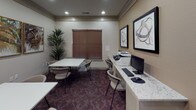 This business center is a well-appointed, single-floor facility designed to support productivity and collaborative work. The space is thoughtfully organized into two functional zones: a dedicated workstation area and an open meeting area.

The workstation area features a granite-topped counter desk equipped with a desktop computer, keyboard, mouse, and a multifunction printer/scanner, providing all the essentials for document management and digital tasks. A tall potted plant beside a window with brown horizontal blinds adds a refreshing, natural touch to this side of the room.

The meeting area accommodates multiple white square tables paired with comfortable cushioned chairs, offering ample space for group discussions or individual work. The patterned deep-burgundy carpet anchors the room with a warm, professional aesthetic.

Décor throughout the space includes framed abstract and nature-inspired artwork, enhancing the visual appeal of the neutral-toned walls. Recessed lighting provides consistent, even illumination across the entire room.

A posted placard on the wall outlines business center rules, and a doorway connects the space to an adjacent area, suggesting convenient access to nearby amenities. The overall ambiance is clean, functional, and professional — well-suited for guests or residents seeking a productive work environment.