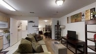 This single-floor residence features an open-concept layout that seamlessly integrates the living, sleeping, and dining areas into one cohesive space. The living area is anchored by a plush sofa adorned with decorative pillows, paired with an upholstered ottoman that doubles as a coffee table, creating a comfortable and inviting lounge zone. A flat-screen television is mounted on a dark wood media console, accompanied by a matching shelving unit that provides organized display and storage space. The sleeping area is thoughtfully partitioned from the main living space using a decorative folding screen, offering a sense of privacy while maintaining the open feel of the floor plan. A floor lamp near the bedside adds warm, ambient lighting to the sleeping nook. The unit features neutral-toned carpeting throughout, white walls, and a textured ceiling with flush-mount light fixtures that provide consistent illumination. Near the entryway, a compact utility area is visible, complete with wall-mounted shelving and decorative accents. Wall art adds a touch of personality to the space. Overall, the layout is efficiently designed to maximize functionality within a compact footprint, making it a practical and comfortable living environment.