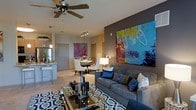 This single-story space showcases an open-concept layout that connects the living, dining, and kitchen areas into one cohesive and functional environment. The living area serves as the focal point, anchored by a plush gray sectional sofa dressed with navy and neutral accent pillows, positioned atop a geometric patterned area rug. A glass-topped coffee table with a metallic frame adds a refined touch to the seating arrangement. Dominating one wall is a large-scale abstract painting in vivid blues, greens, and warm tones, mounted against a contrasting dark accent wall that adds dramatic depth to the room. A ceiling fan with light fixture overhead provides both comfort and illumination. The dining area, positioned adjacent to the living space, features a round table with upholstered white chairs and is adorned with its own bold artwork. Track lighting highlights the dining zone effectively. The kitchen, visible beyond, offers stainless steel appliances, sleek cabinetry, and pendant lighting. Neutral-toned walls and light carpeting throughout create a warm, welcoming backdrop that allows the colorful décor and artwork to take center stage.