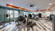 Located at 1325 Boland Pl in Richmond Heights, Missouri, this fitness center offers a spacious and thoughtfully designed workout environment across a single floor. The facility comprises two exercise rooms that together provide a comprehensive range of training options for users of all fitness levels.

The interior design is visually striking, with a bold orange accent band running along the upper perimeter of the walls, contrasting against teal-blue lower walls and a crisp white ceiling. Recessed lighting and ceiling fans ensure the space remains well-lit and comfortable during workouts. Full-length mirrored walls on one side visually expand the rooms while allowing exercisers to monitor their form.

The flooring features a modern geometric pattern in neutral tones, combining light wood-style planks with contrasting dark accents, adding a dynamic aesthetic to the space. Large windows along one wall invite abundant natural light and offer views of an outdoor pool and surrounding grounds, creating an energizing atmosphere.

The facility is comprehensively equipped with TRUE-brand cardio machines, including treadmills, ellipticals, stair climbers, and a rowing machine. A dedicated free weights area includes a full dumbbell rack and weight benches, while additional strength training machines line the walls. Wall-mounted flat-screen televisions are positioned throughout for entertainment during workouts, rounding out this full-featured fitness destination.