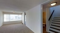 This two-floor residential space offers a straightforward and functional layout designed for comfortable everyday living. The main living area features soft wall-to-wall carpeting underfoot, creating a warm and inviting atmosphere. A large window floods the space with natural light, offering views of the surrounding exterior and enhancing the open feel of the room. The textured ceiling adds a classic touch to the interior finish throughout.

A partition wall defines the transition between the living area and an adjoining bathroom, which is conveniently accessible from the main floor. The bathroom features hardwood-style flooring, providing a clean contrast to the carpeted living spaces. The layout is thoughtfully arranged to ensure ease of movement between the key areas of the home.

Clean white walls throughout the space lend a neutral, versatile backdrop that accommodates a variety of interior styles and furnishing choices. The overall ambiance is bright, practical, and well-organized, with a clear separation of living and utility spaces across the two floors. This space is well-suited for those seeking a functional home with a simple, clean aesthetic.