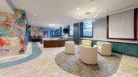 Located on Euclid Avenue in the heart of Cleveland, Ohio, this thoughtfully designed single-floor communal space offers a seamless blend of modern aesthetics and functional layouts. The open floor plan is anchored by two distinct living areas, each with its own character. The primary lounge features curved sectional seating, cream-toned swivel chairs, and layered geometric carpeting, all illuminated by dramatic gold-rimmed circular pendant lights. A wall-mounted television is framed by deep teal tile accents and wood-trimmed windows that invite natural light. A secondary lounge area offers modular periwinkle sofas, intimate booth-style seating nooks with gradient blue walls, and a relaxed reading corner complete with a coffee table. A vibrant large-scale abstract mural and an upright piano add artistic character near the entry corridor, which features rich wood-and-frosted-glass doors. Both kitchens are elegantly appointed with teal shaker cabinetry, marble-look quartz countertops, white geometric tile backsplashes, gold hardware fixtures, and striking globe chandelier lighting. The herringbone-patterned flooring flows consistently throughout, tying together the entire space with warmth and cohesion.