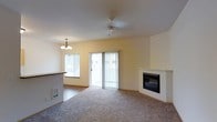 This single-floor residence offers a welcoming and functional layout designed for comfortable everyday living. The main living area is carpeted and bathed in natural light, courtesy of a large sliding glass door fitted with vertical blinds that leads to an outdoor area, as well as an adjacent window that further brightens the space. A classic white-mantled fireplace anchors one corner of the living room, adding a warm, inviting focal point to the room.

A ceiling fan is centrally positioned overhead, ensuring year-round comfort throughout the open space. The living area flows seamlessly into a dining space, which is distinguished by a decorative chandelier that adds a touch of elegance. A raised counter or breakfast bar separates the dining area from what appears to be an adjoining kitchen, offering a convenient and social layout ideal for both casual meals and entertaining.

The walls are finished in a soft, neutral tone that complements the light-colored carpeting, creating a clean and airy ambiance throughout. A floor-level vent is visible along the baseboard, indicating a built-in climate control system. Overall, this single-floor space combines practical design with comfortable features, making it a well-appointed and livable home.